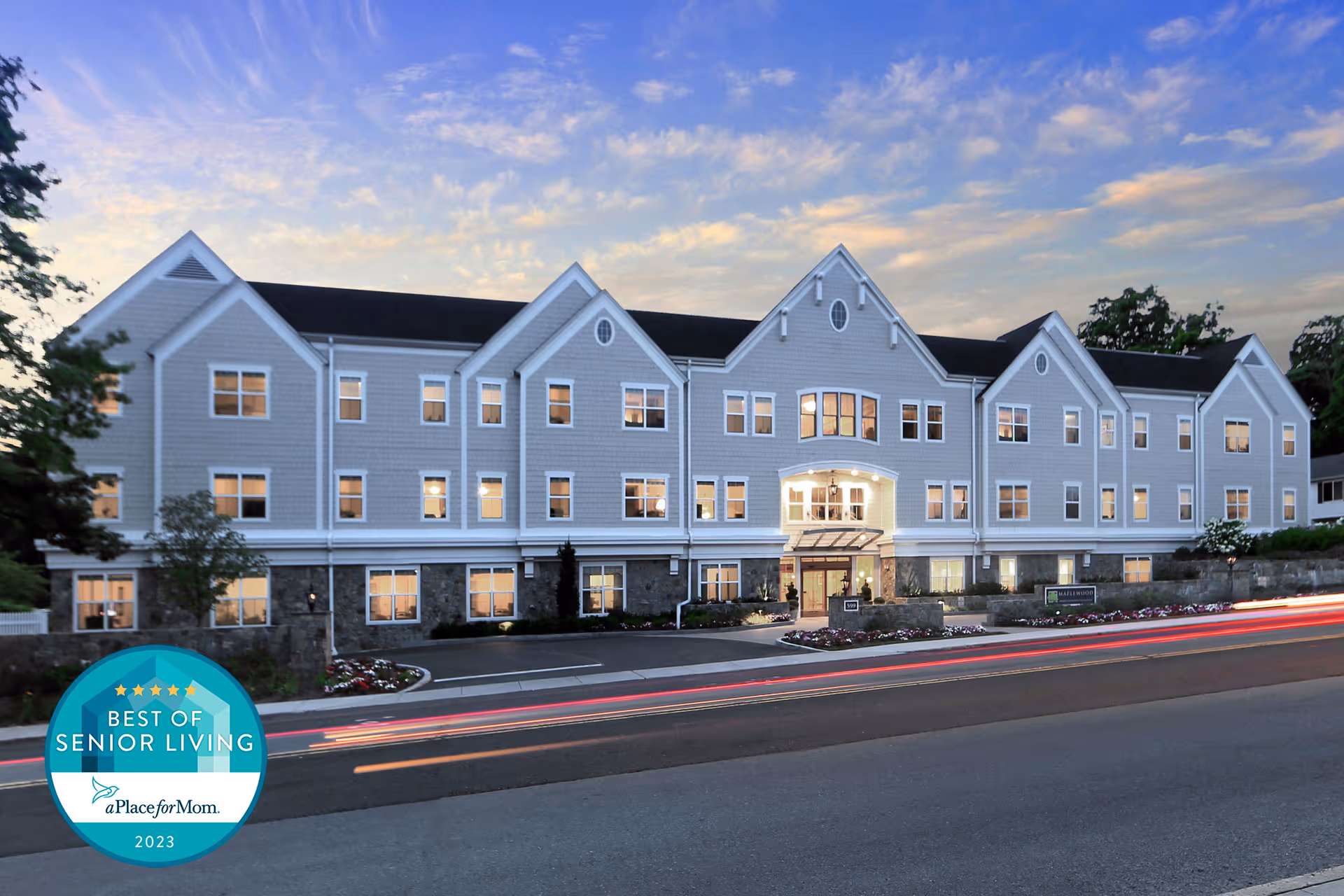 Exterior view of a large, three-story senior living facility building named Maplewood at Darien during dusk with lights on inside. The building has a gray facade with white trim and multiple windows. There is a driveway and landscaped flower beds in front, with a partly cloudy sky in the background.