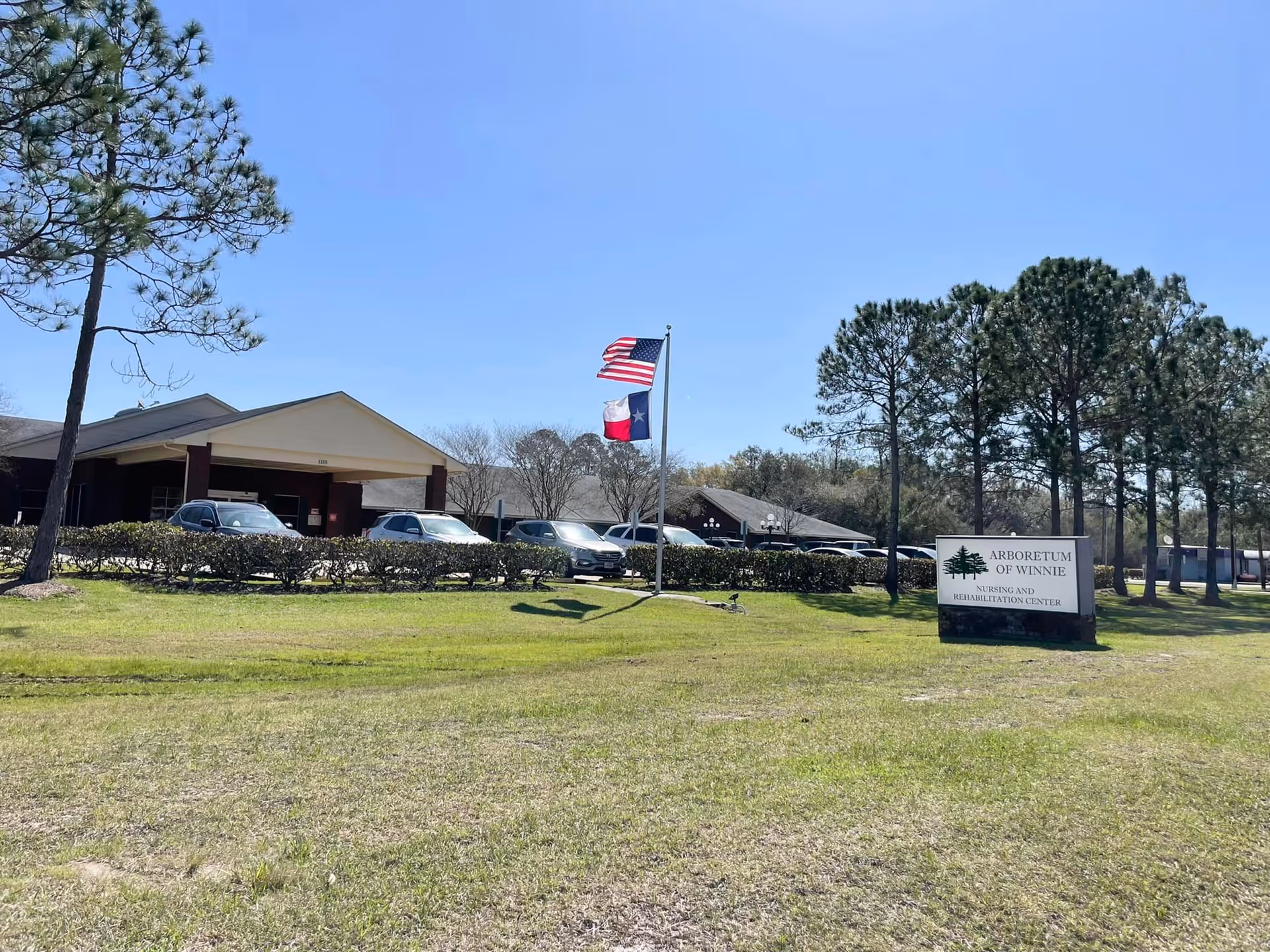 Front view of Arboretum of Winnie nursing and rehabilitation center showing the building entrance, flagpoles, and a sign on the lawn.
