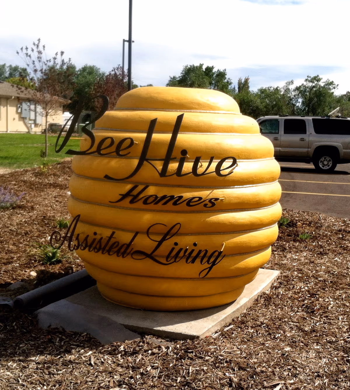 A large yellow beehive-shaped sign with black cursive text that reads 'BeeHive Homes Assisted Living' situated outdoors on a mulch bed with a parking lot and trees in the background.