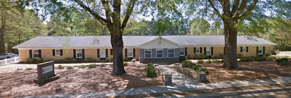 Single-story senior living facility building with beige exterior walls, multiple windows, and a gray roof. There are two large trees in front of the building, a paved walkway leading to the entrance, and landscaped areas with mulch and shrubs. A sign is visible near the left side of the walkway.