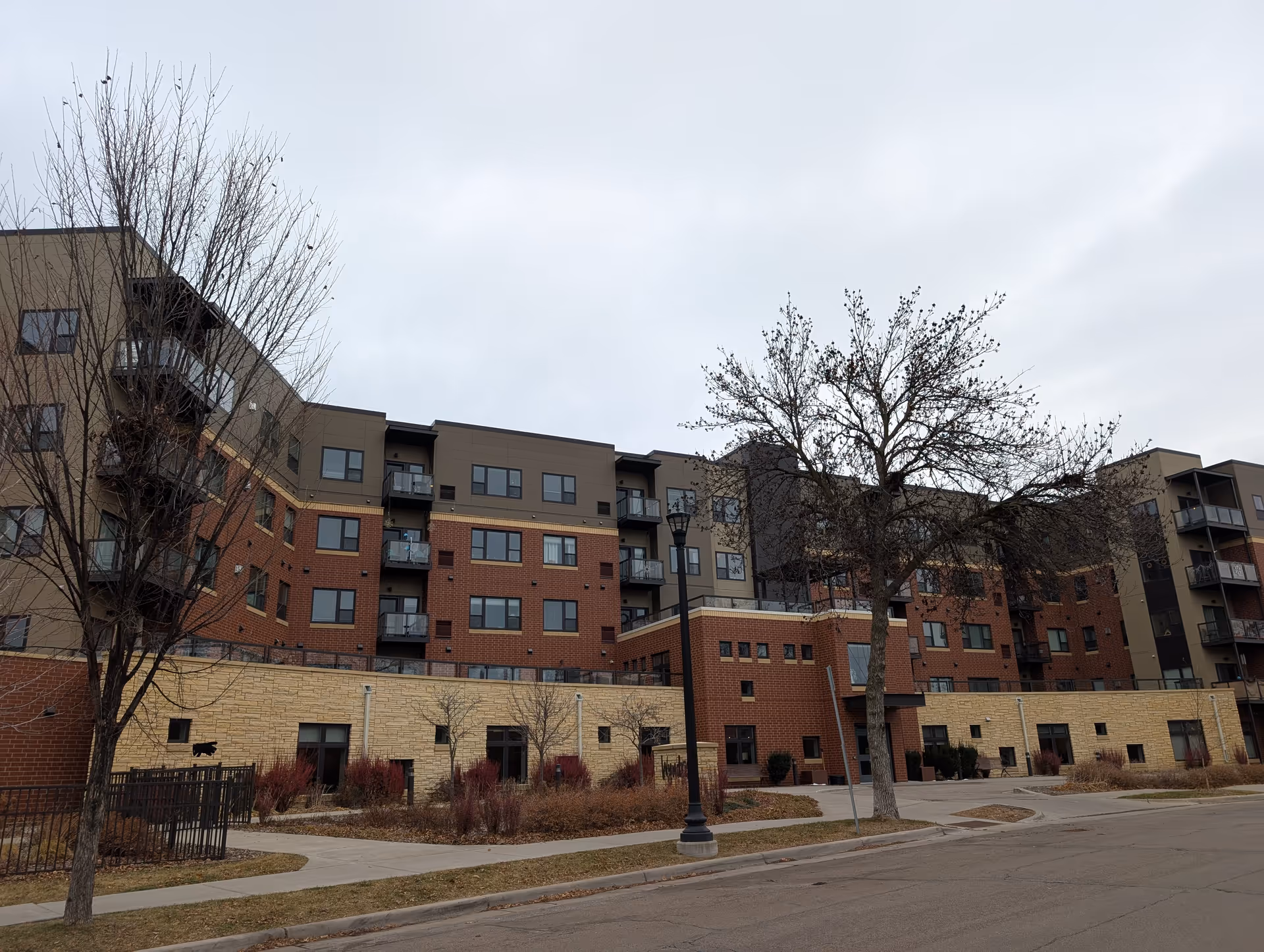 Exterior front of a multi-story senior living building with brick and stone facade, balconies, leafless trees, and a sidewalk.