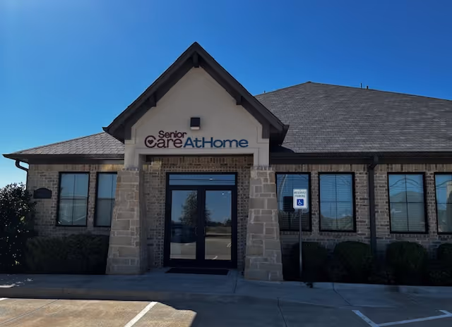 Front exterior of Senior Care At Home building with entrance, stone pillars, and parking.