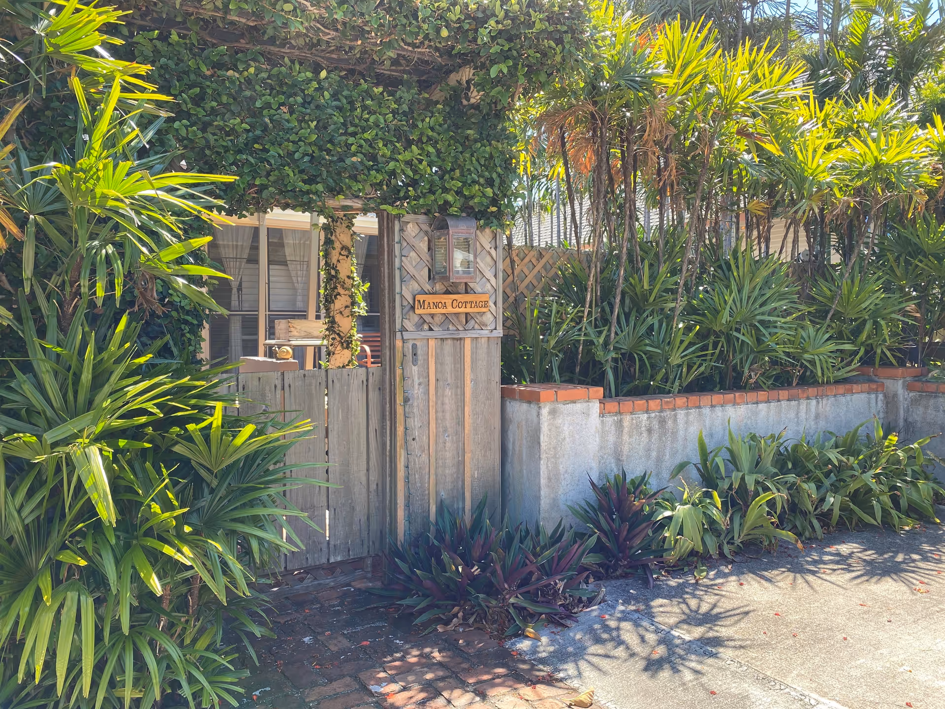 Wooden gate labeled Manoa Cottage, surrounded by lush tropical plants at entrance.