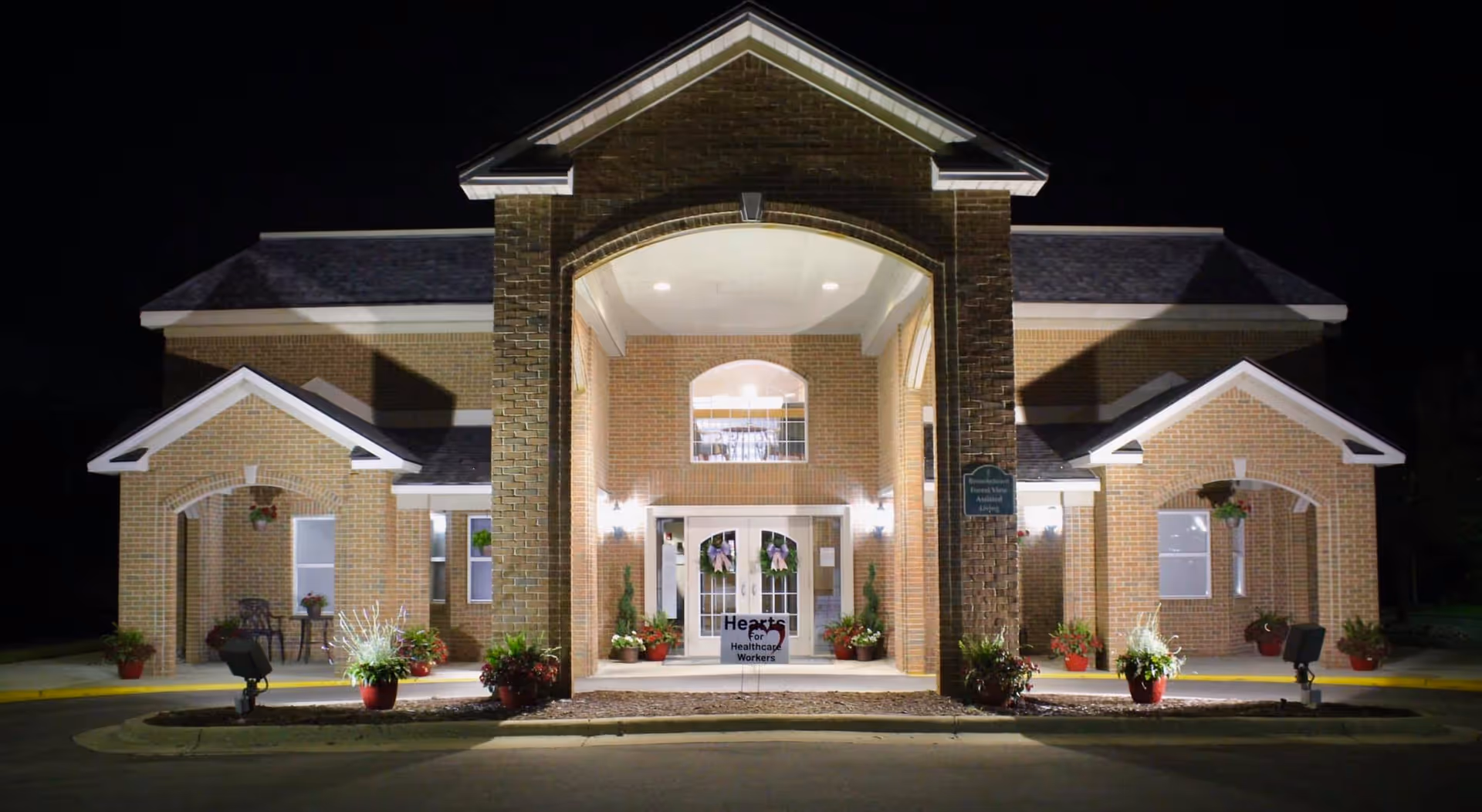 Night view of Brownstown Forest View Assisted Living entrance with brick facade and plants.