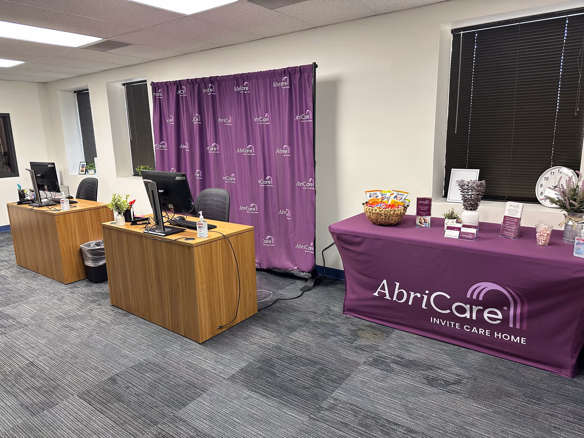 Open office area with two desks, computers, and a purple AbriCare event table.