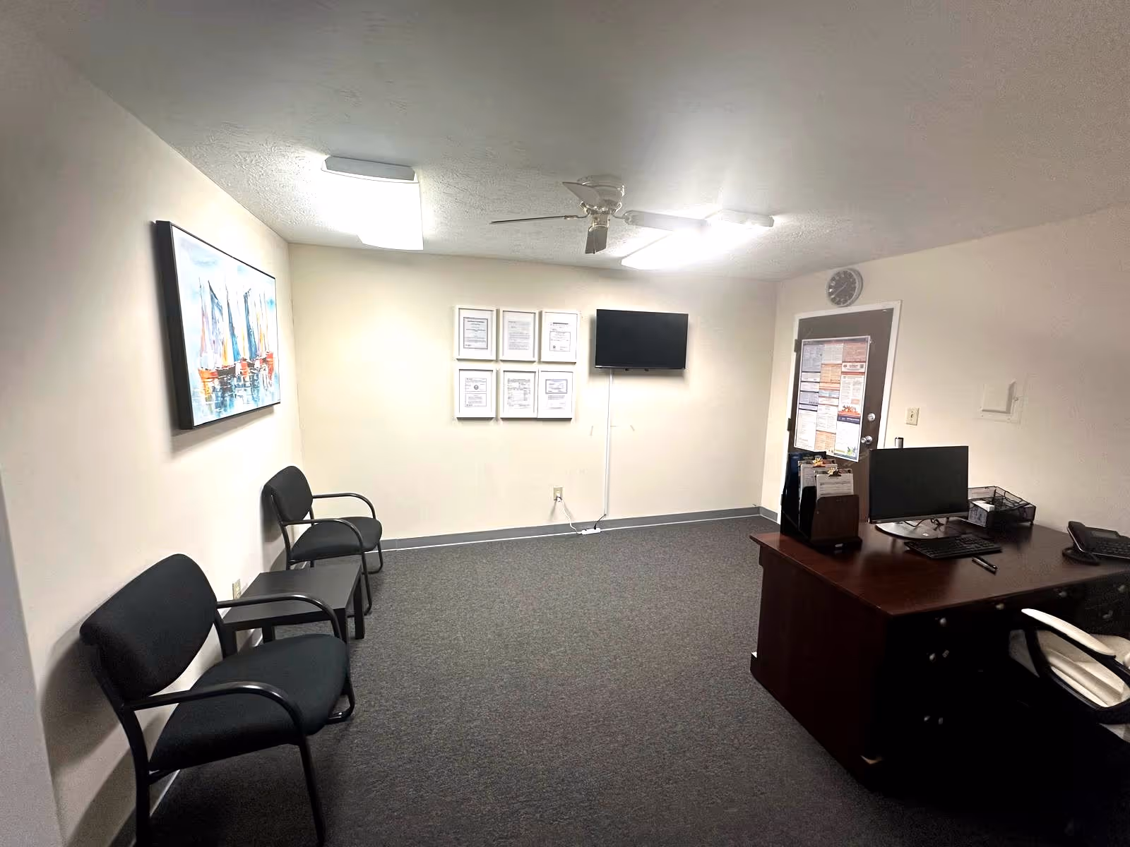 Reception area with desk, computer, wall certificates, mounted TV, and waiting chairs.