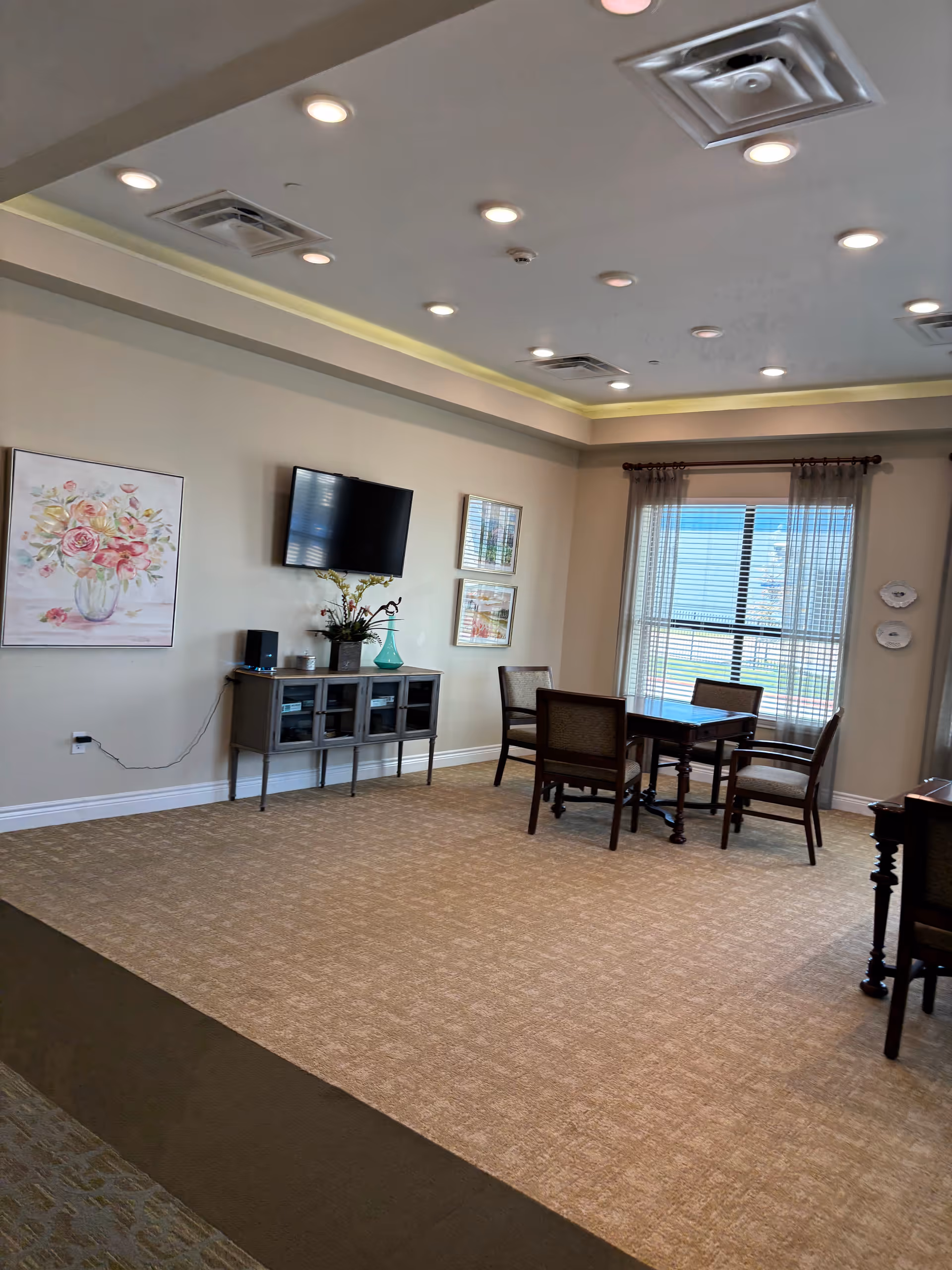 Bright senior living lounge with TV, artwork, seating area, and large windows.