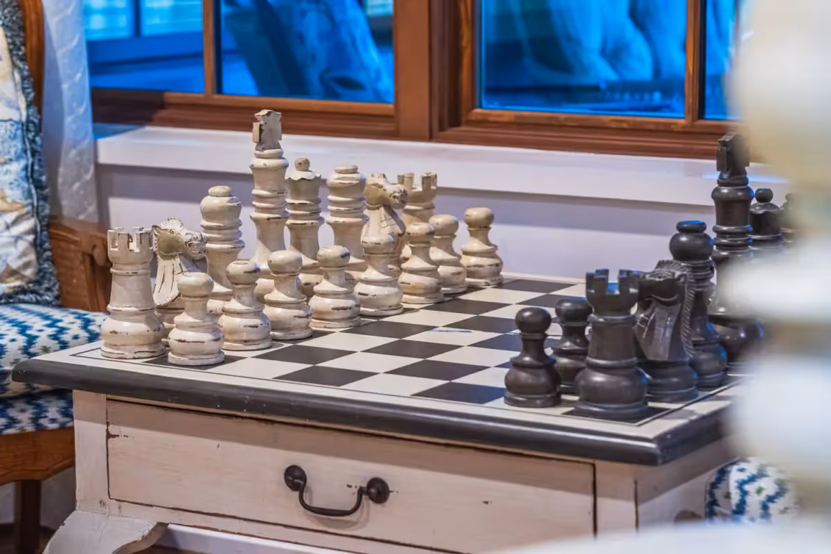 A chessboard with oversized white and black pieces sits on a wooden table inside a cozy indoor room.