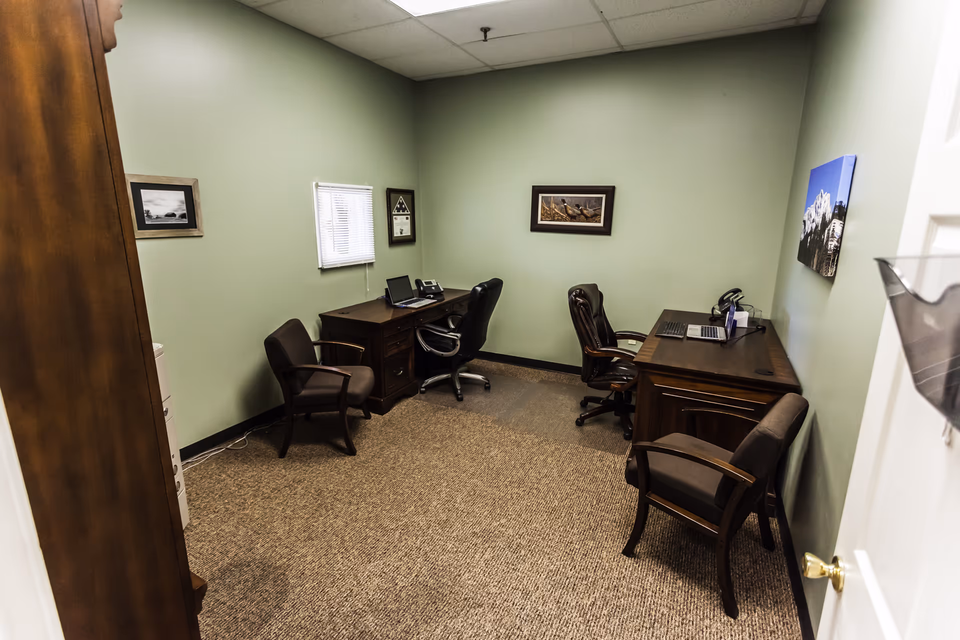 Two dark wooden desks with laptops and chairs in a green-walled office.