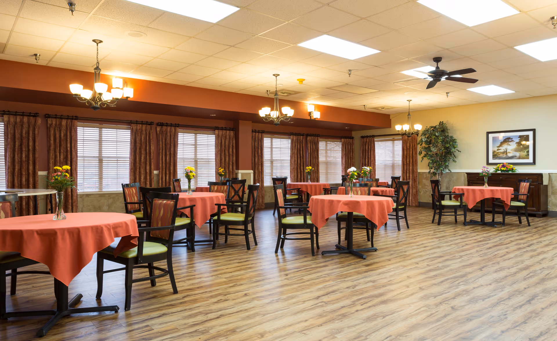 A spacious dining room with several round tables covered with red tablecloths, each table surrounded by dark wooden chairs with green cushions. The room has large windows with brown curtains, wooden flooring, chandeliers hanging from the ceiling, and a ceiling fan. There are flower vases on each table and a framed picture on the far wall next to a large potted plant.