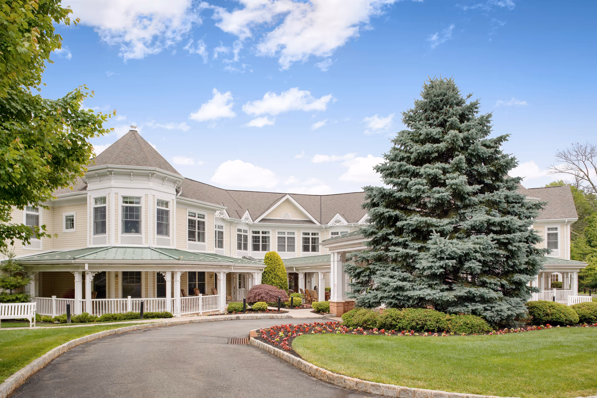 Exterior view of a senior living facility with a circular driveway, well-maintained lawn, colorful flower beds, and a large evergreen tree in front. The building is two stories with multiple windows and a covered porch area.