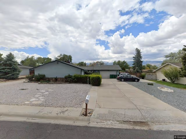 Single-story residential building with a large concrete driveway and gravel landscaping in the front yard. There is a black car parked on the right side of the driveway. The sky is partly cloudy with some blue patches visible.