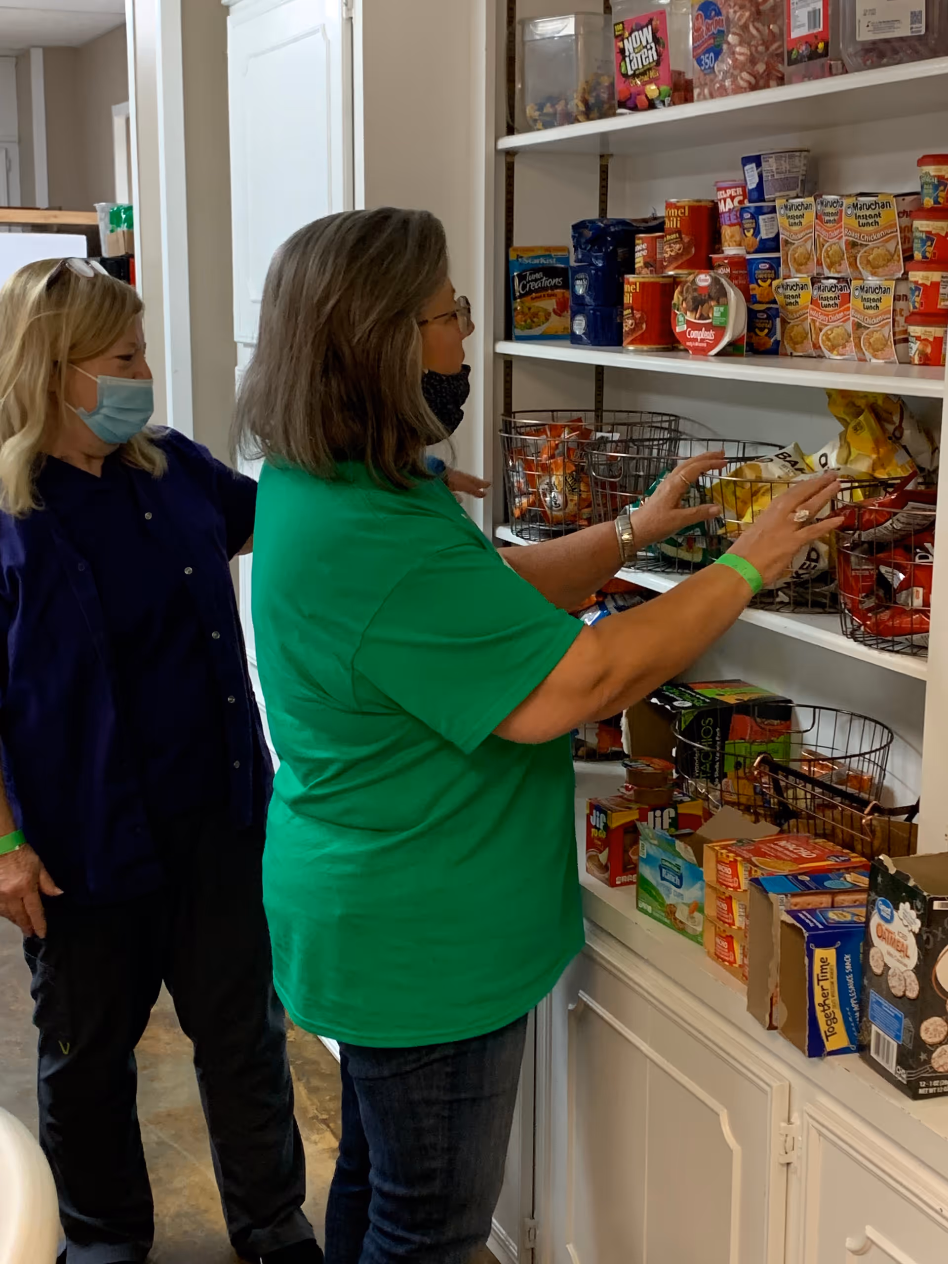 Two women wearing face masks stand in front of shelves stocked with various packaged food items. One woman in a green shirt is reaching for a bag of chips, while the other woman in a dark blue shirt stands behind her. The shelves contain canned goods, snacks, and boxed food items.