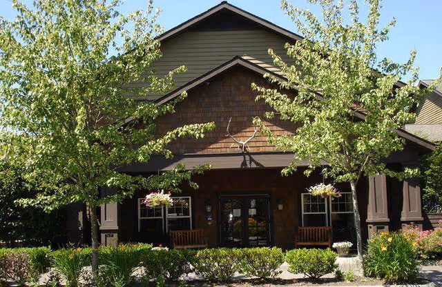Front exterior view of a building with a peaked roof, brown wooden siding, and a covered porch area with benches. The entrance is flanked by two windows and surrounded by green trees and shrubs under a clear blue sky.