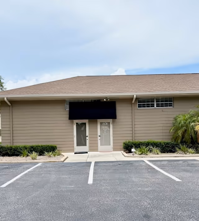 Front exterior of a single-story facility with double doors, a black sign, and parking.