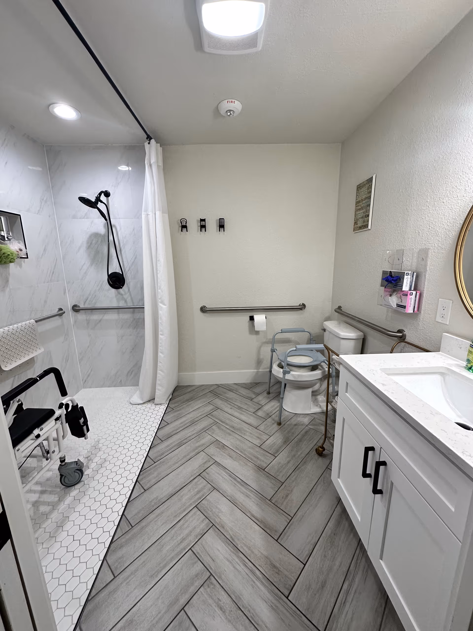 A clean, accessible bathroom featuring a walk-in shower with a white curtain, a black showerhead, and a shower chair. The floor has a herringbone pattern with gray tiles. There is a toilet with a raised seat and grab bars on the walls for support. A white vanity with a sink and a round mirror is visible on the right side. The bathroom is well-lit with ceiling lights.