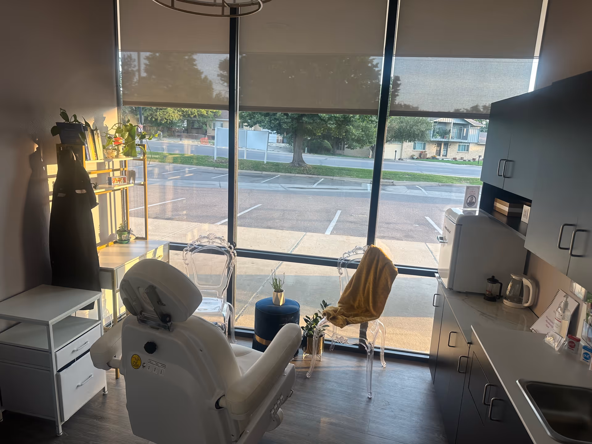 A dental chair in a bright clinic room with cabinets, a window, and small seating.