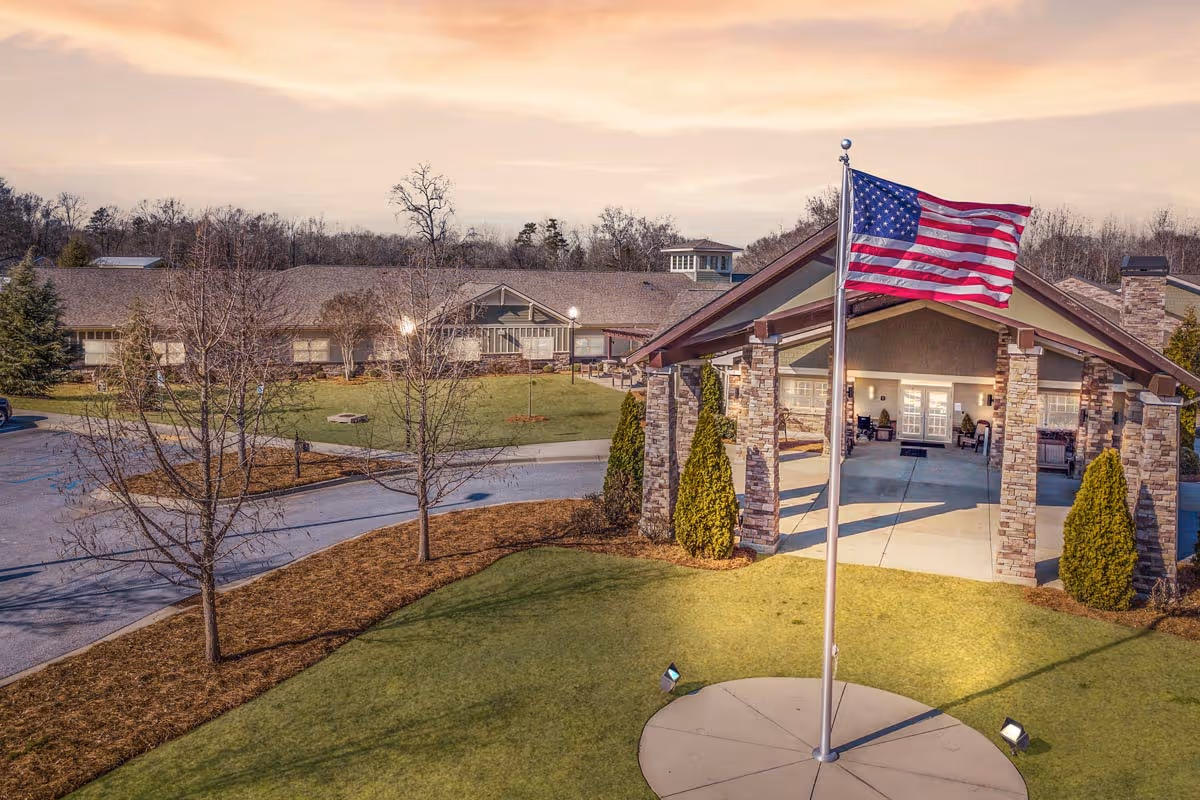 Exterior view of a senior living facility with a large covered entrance supported by stone pillars, an American flag on a flagpole in the foreground, and a well-maintained lawn and trees surrounding the building under a cloudy sky.