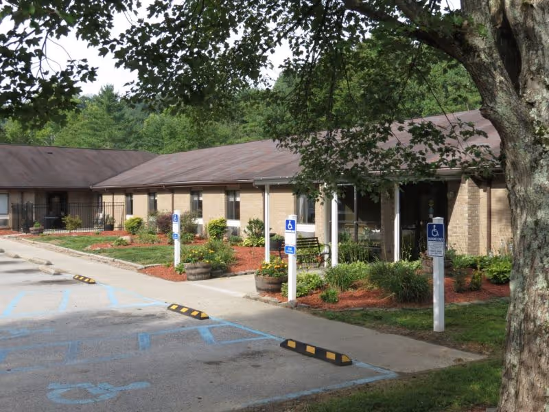 Front exterior of a single-story brick senior care building with accessible parking spaces, landscaped flower beds, and a large tree in the foreground.