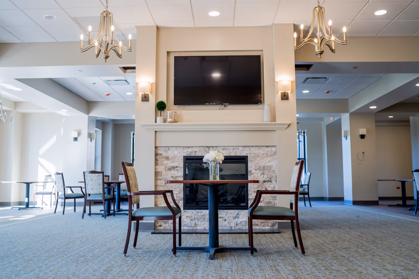 A bright communal dining and lounge area with a central fireplace and wall-mounted TV, small tables and chairs, and chandeliers.