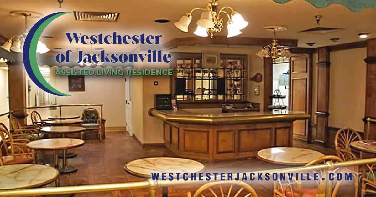 Interior view of a common area in Westchester of Jacksonville Assisted Living Residence featuring several round tables with chairs, a wooden reception or service counter, warm lighting fixtures on the ceiling, and wood-paneled walls.