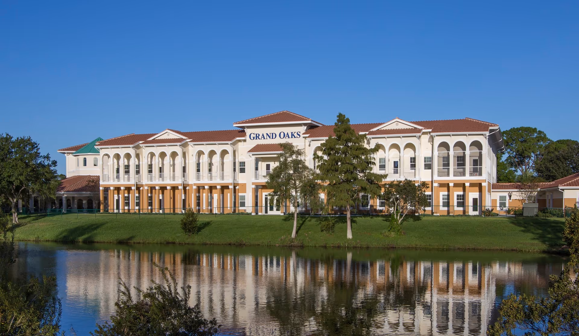 Exterior view of the Grand Oaks senior living facility building with a clear blue sky and a pond reflecting the building in the foreground.