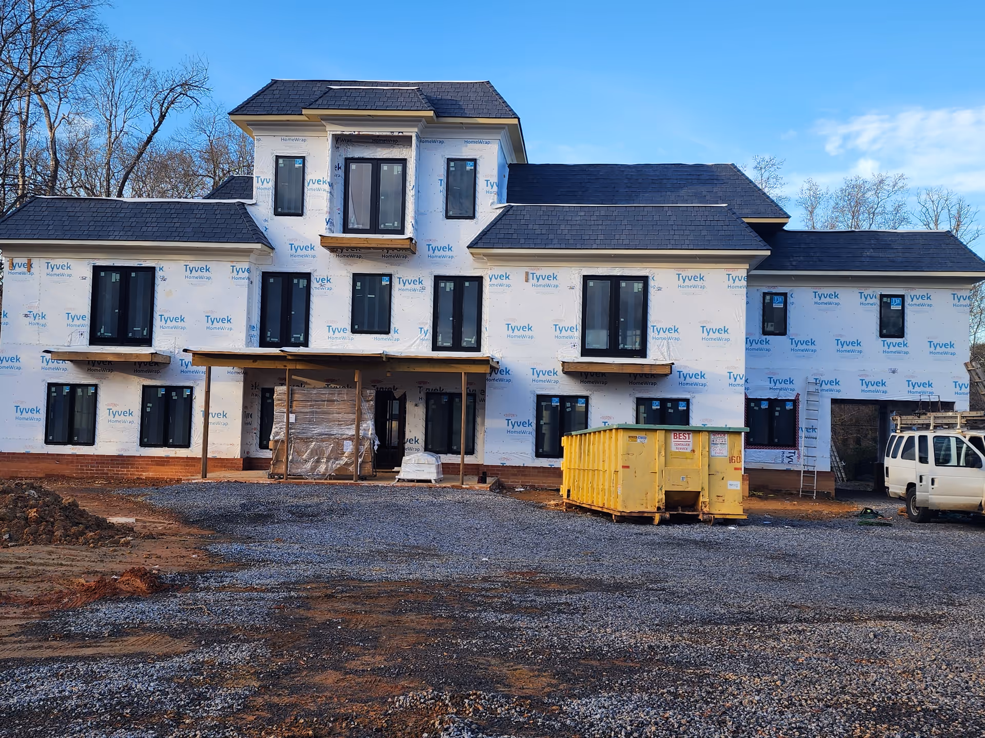Under construction two-story building wrapped in Tyvek with a yellow dumpster in a gravel lot.