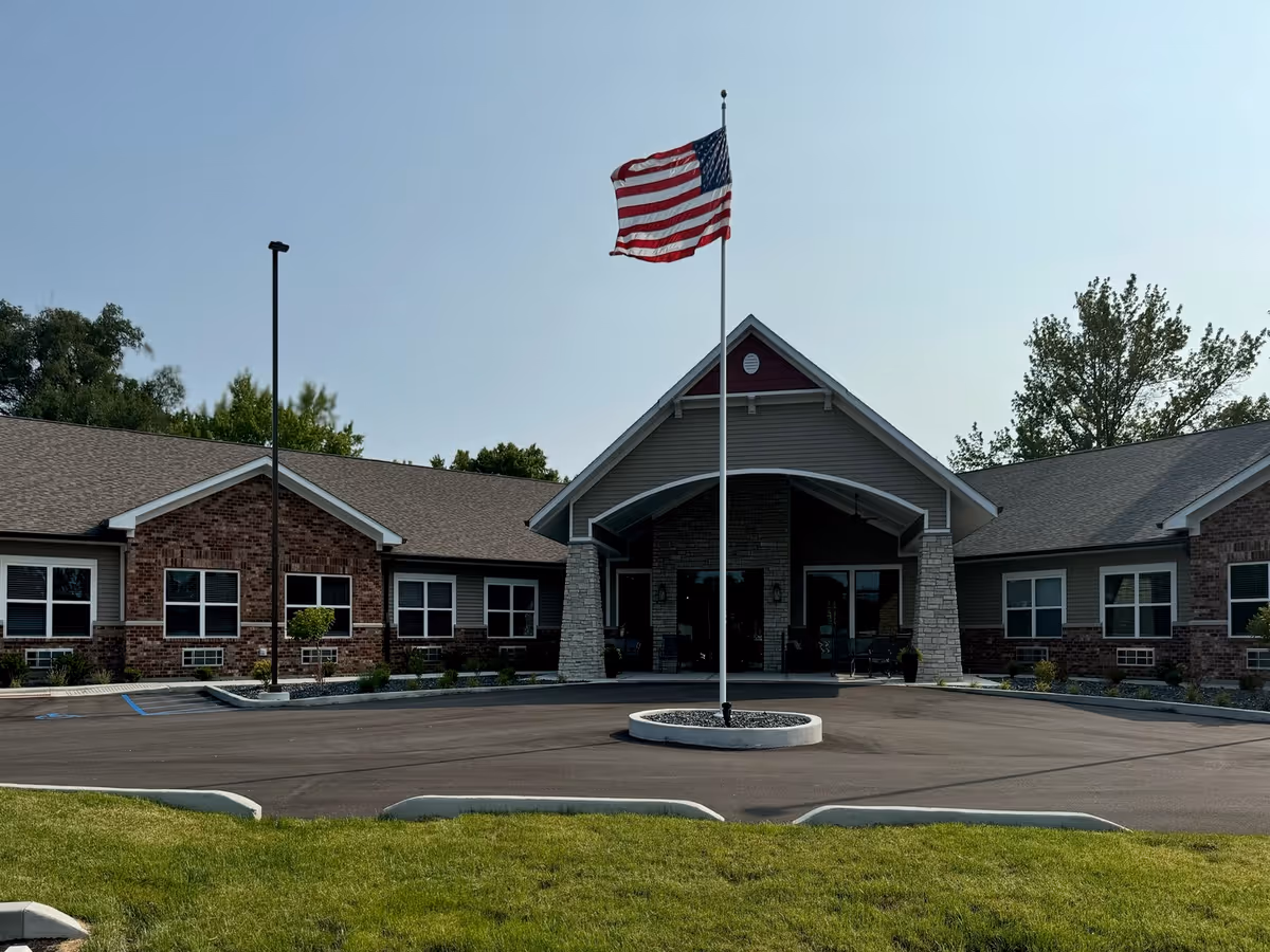 Front exterior view of Autumn Trace Kentland Assisted Living facility with a large American flag on a flagpole in the center of a circular driveway. The building has a brick and siding facade with multiple windows and a covered entrance. Trees and greenery are visible in the background under a clear sky.