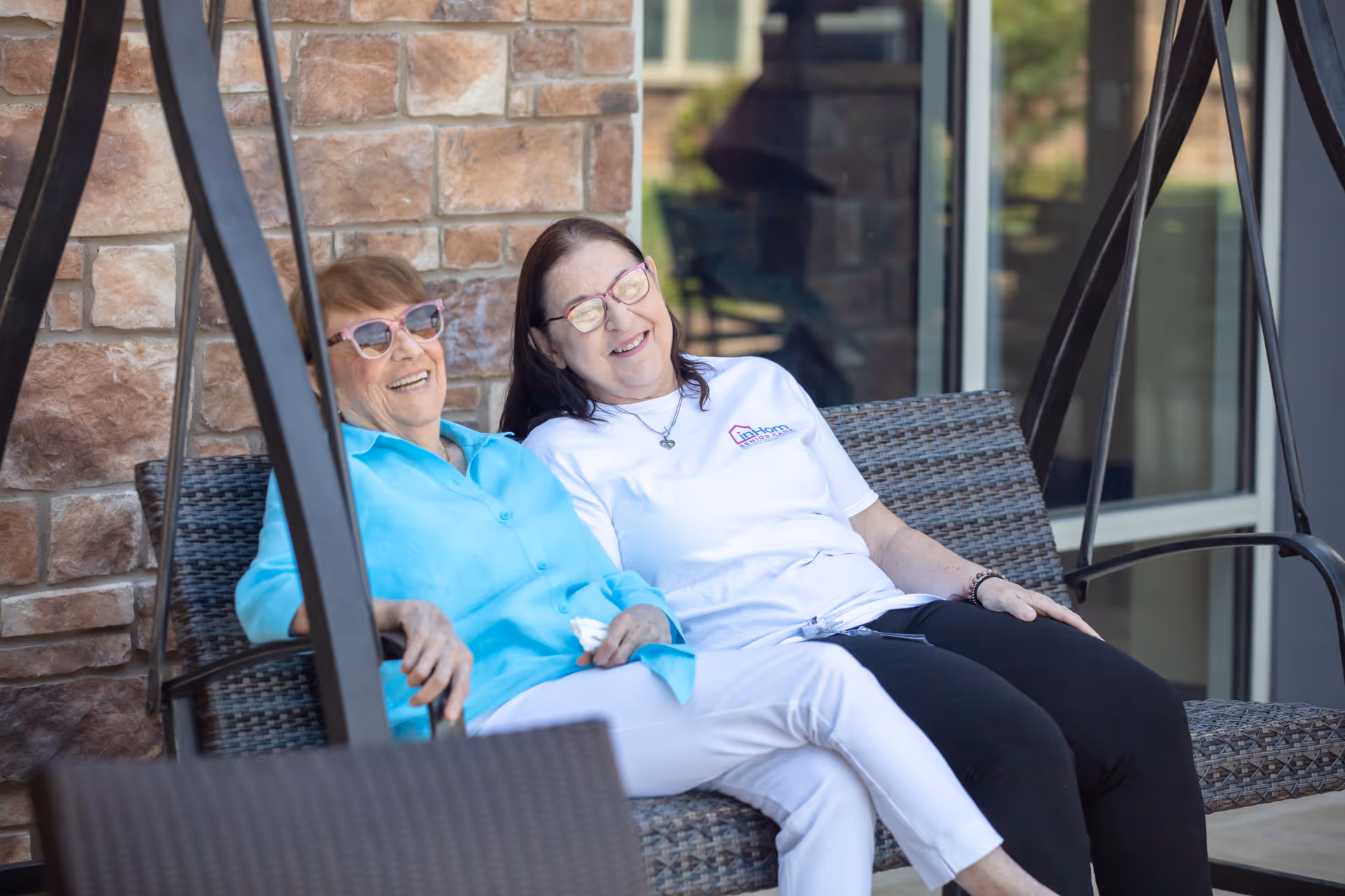 Two elderly women sit on a wicker outdoor swing, smiling beside a brick wall.
