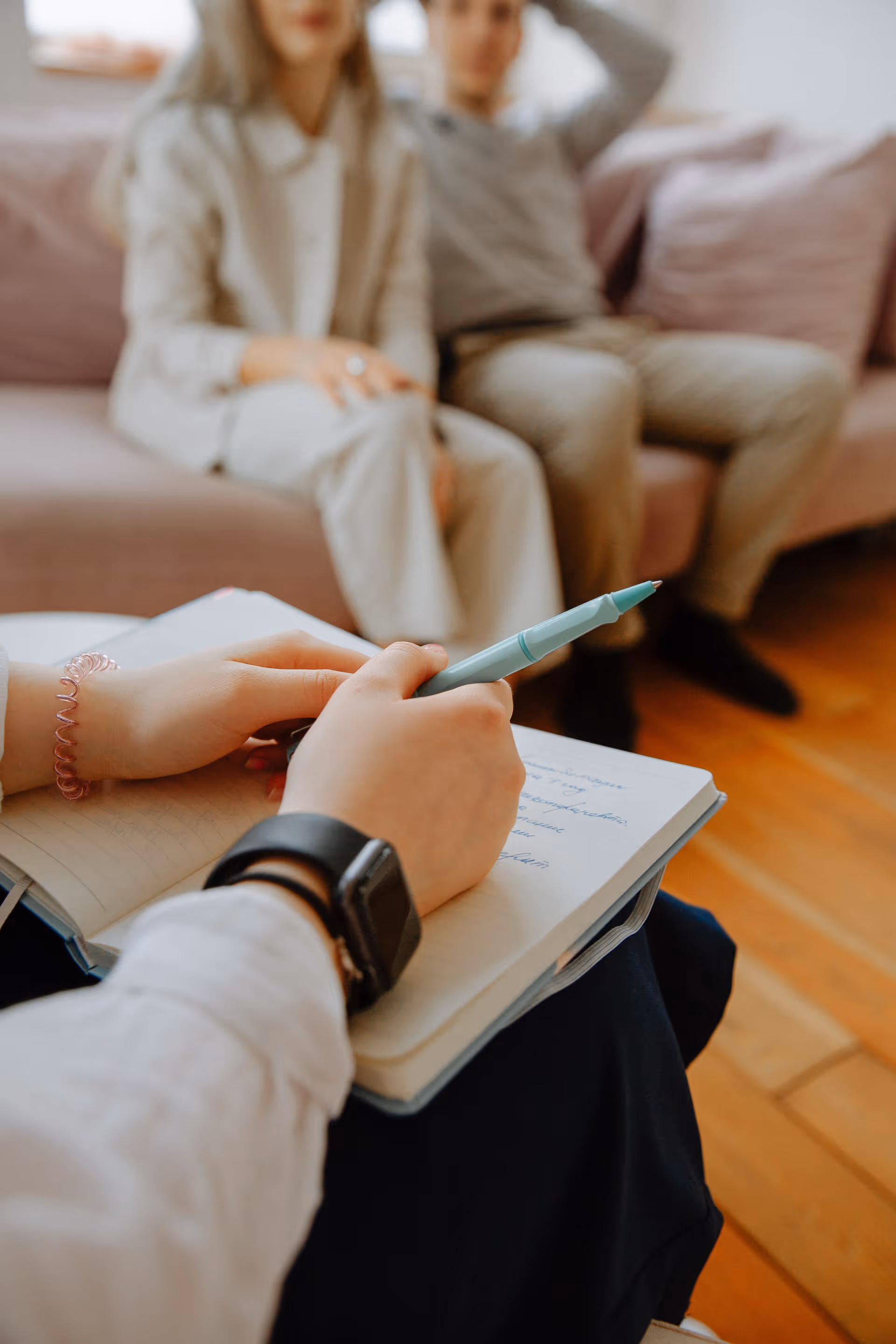 A person writes in a notebook on their lap while two residents sit on a couch.