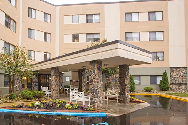Entrance area of a multi-story senior living facility with stone pillars supporting a covered drop-off area. White benches are placed under the covered area, and there are landscaped flower beds and small trees around the entrance. The building has beige and light brown exterior walls with multiple windows.