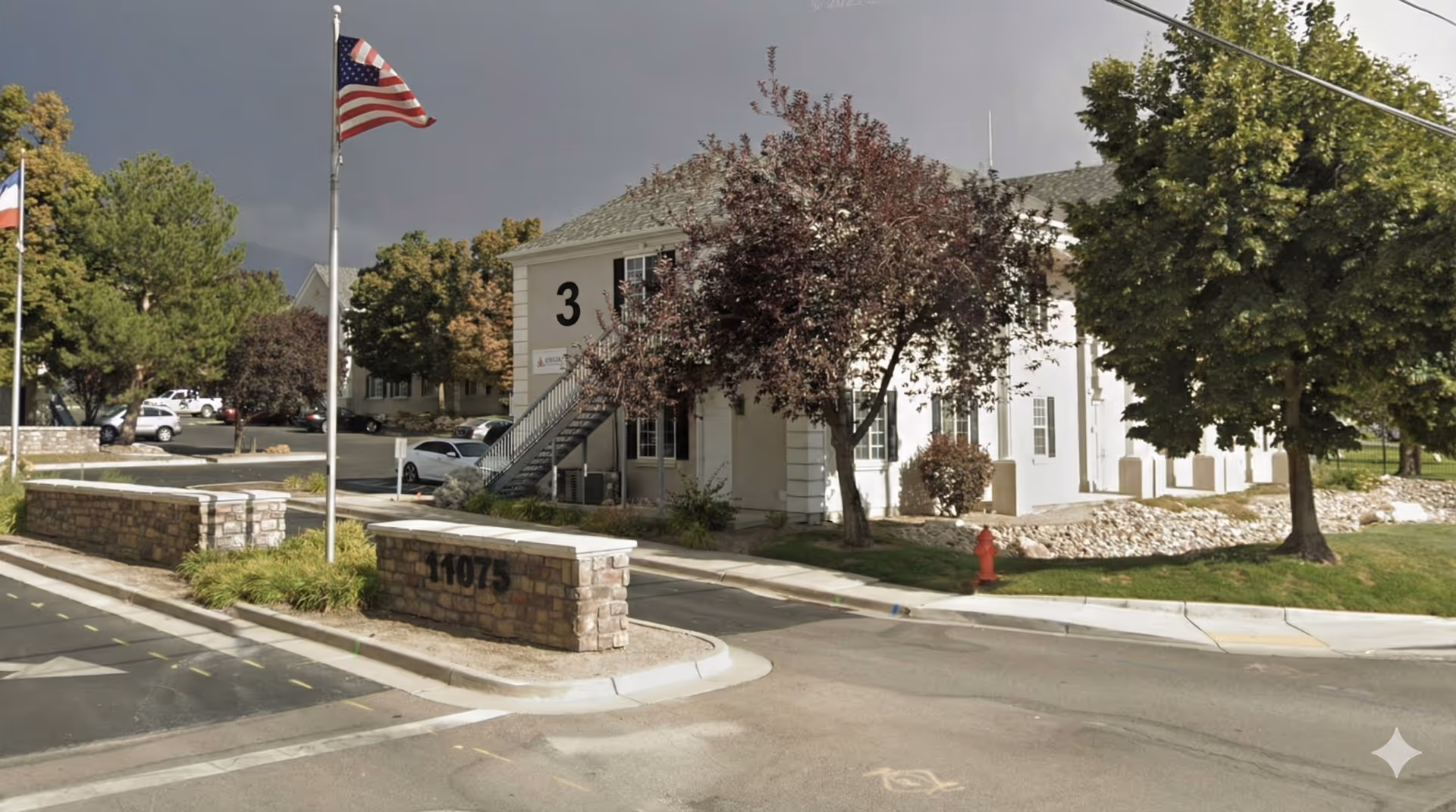 White two-story senior living building with stairs, flagpoles, trees, and a stone entry wall.