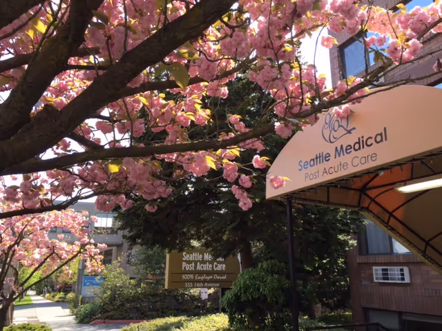 View of the exterior entrance area of Seattle Medical Post Acute Care facility with blooming pink cherry blossom trees in the foreground and signage displaying the facility's name and address.