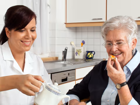 A caregiver in a white uniform pours tea from a teapot while an elderly woman with gray hair and glasses smiles and holds a small pastry in a kitchen setting.
