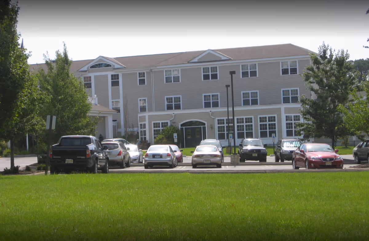 Three-story senior living facility with beige siding, parking lot, and green lawn.