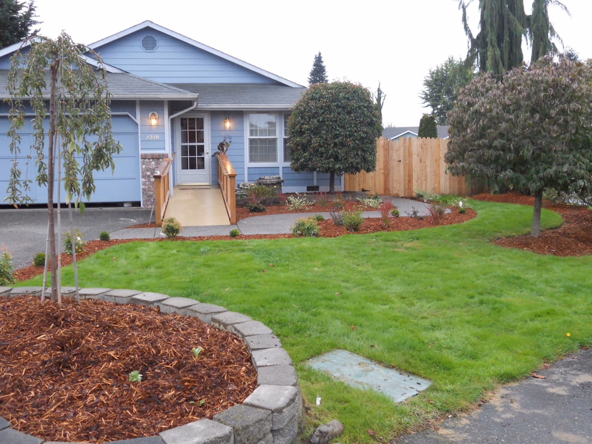 Front yard and entrance of a blue single-story house with a ramp, lawn, and landscaped beds.