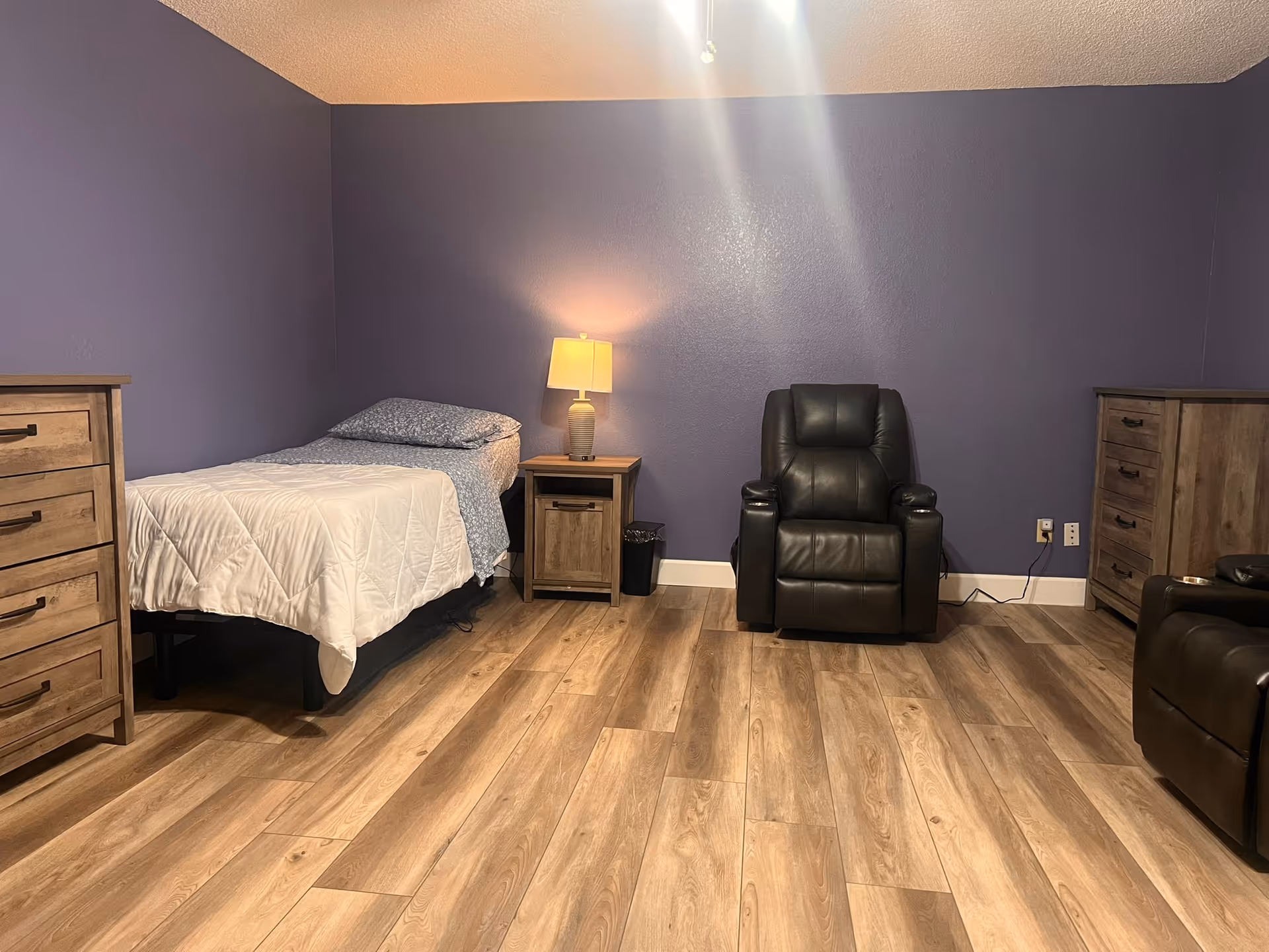 A bedroom in a senior care facility with a bed, recliner, and wooden dressers.