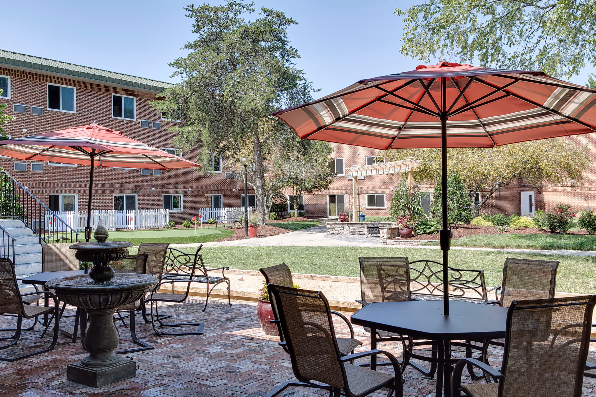 Outdoor patio area with tables and chairs under large red umbrellas, a brick-paved floor, a water fountain, and a grassy courtyard with trees and a brick building in the background.
