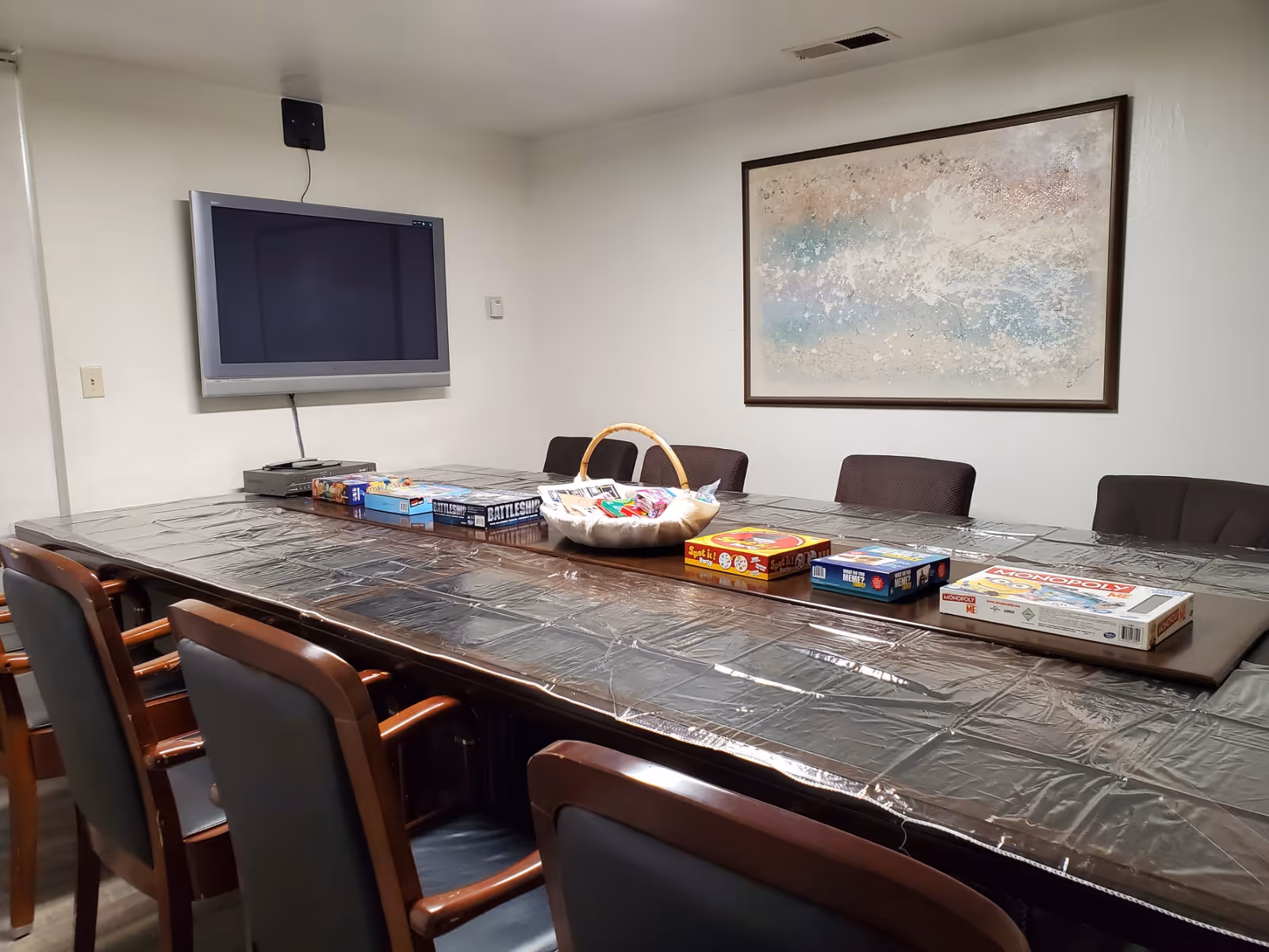 A conference or activity room with a long table covered in a plastic tablecloth. Several board games are arranged on the table including Monopoly, Battleship, and Spot It. There are multiple chairs around the table, a wall-mounted flat screen TV, and a large abstract painting on the wall.