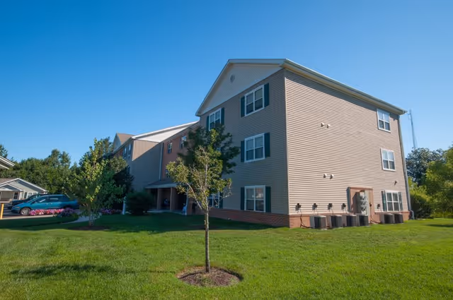 Three-story beige residential building with green shutters on a grassy lawn and small trees under a clear blue sky.