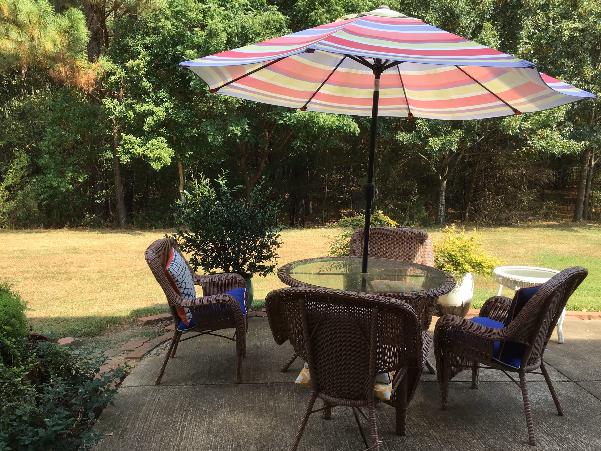 Outdoor patio area with a round glass-top table surrounded by four wicker chairs, each with cushions. A large striped umbrella provides shade over the table. The patio overlooks a grassy yard with trees and shrubs in the background.