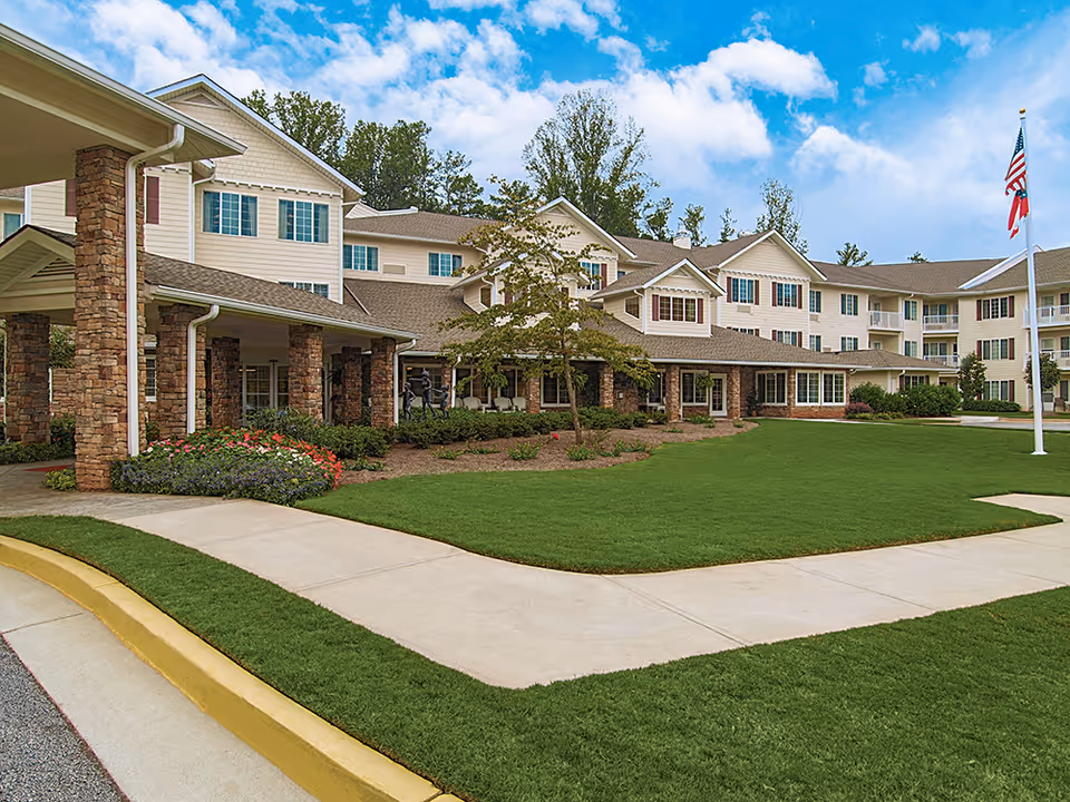 Front exterior of a multi-story senior living building with stone columns, a covered entrance, manicured lawn, walkway, and a flagpole.
