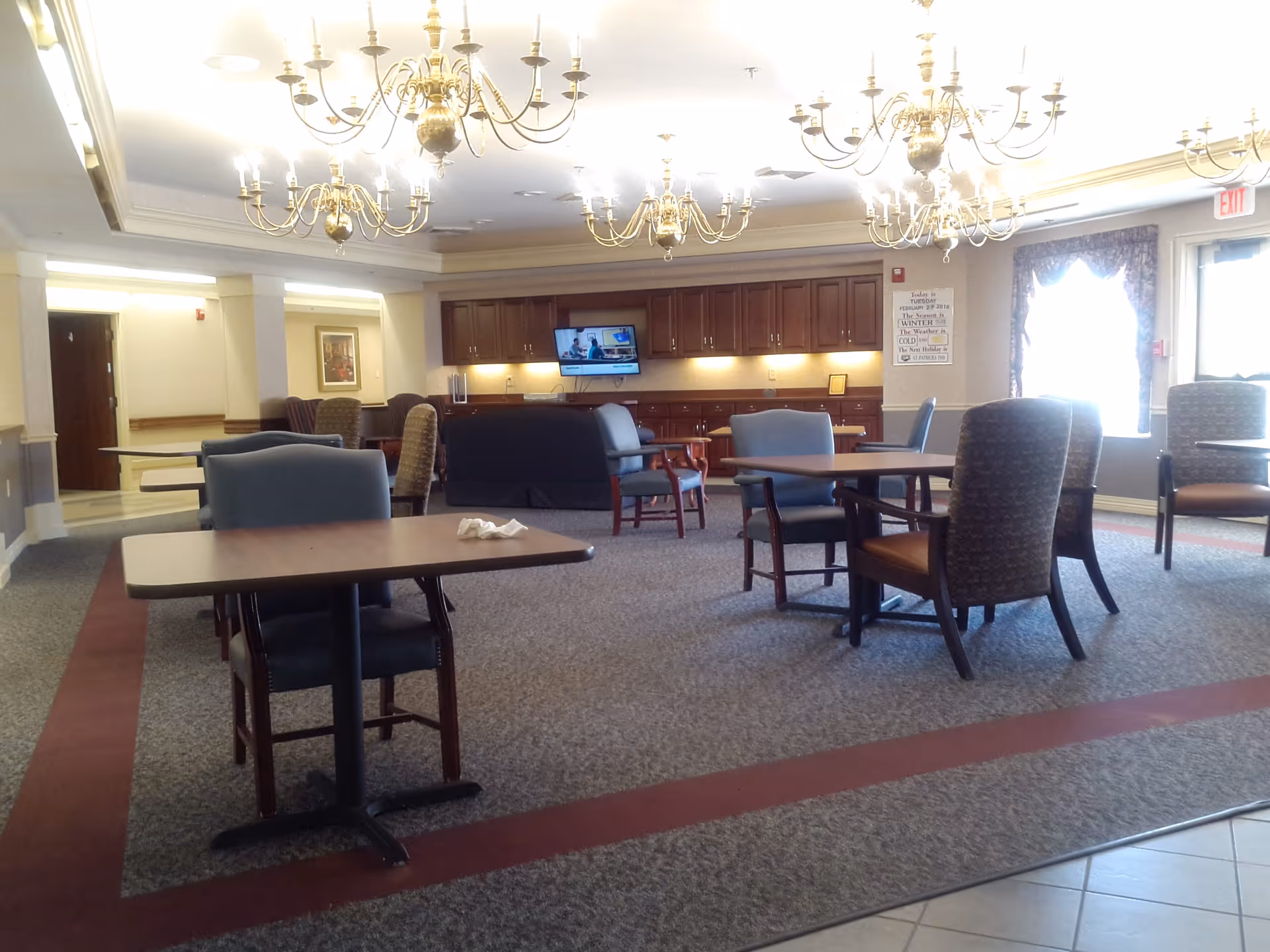 Interior senior living common area with chandeliers, tables, chairs, and a mounted TV.