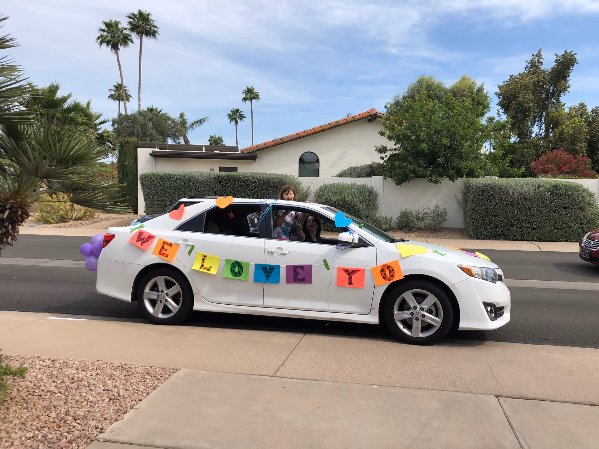 A white car decorated with rainbow banner and balloons drives past a single-story assisted living home.