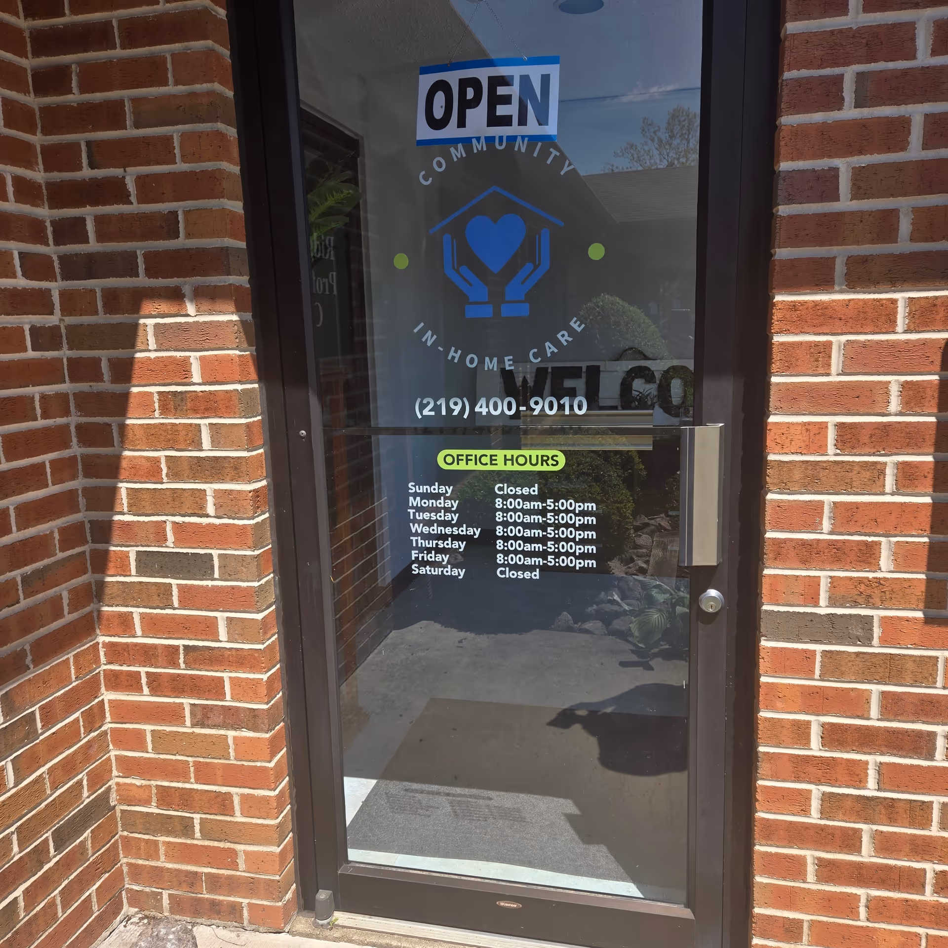 Brick building entrance with glass door displaying an OPEN sign and office hours.