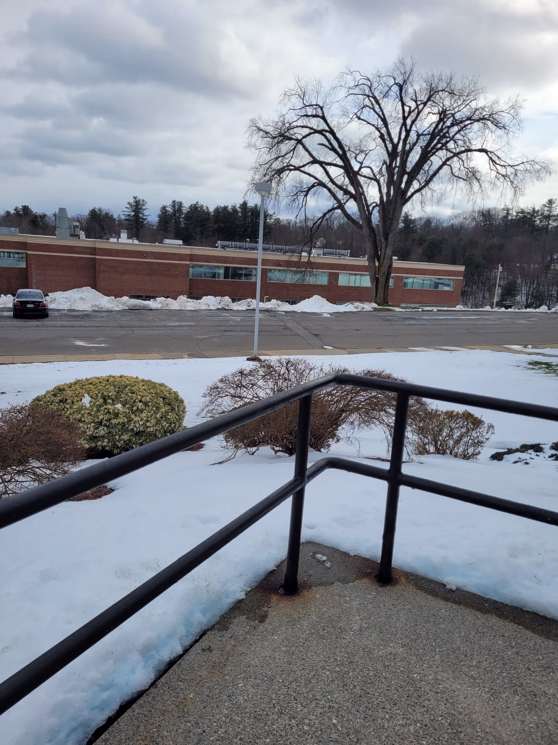 Snow-covered outdoor railing in foreground, leafless tree and brick facility building in distance.