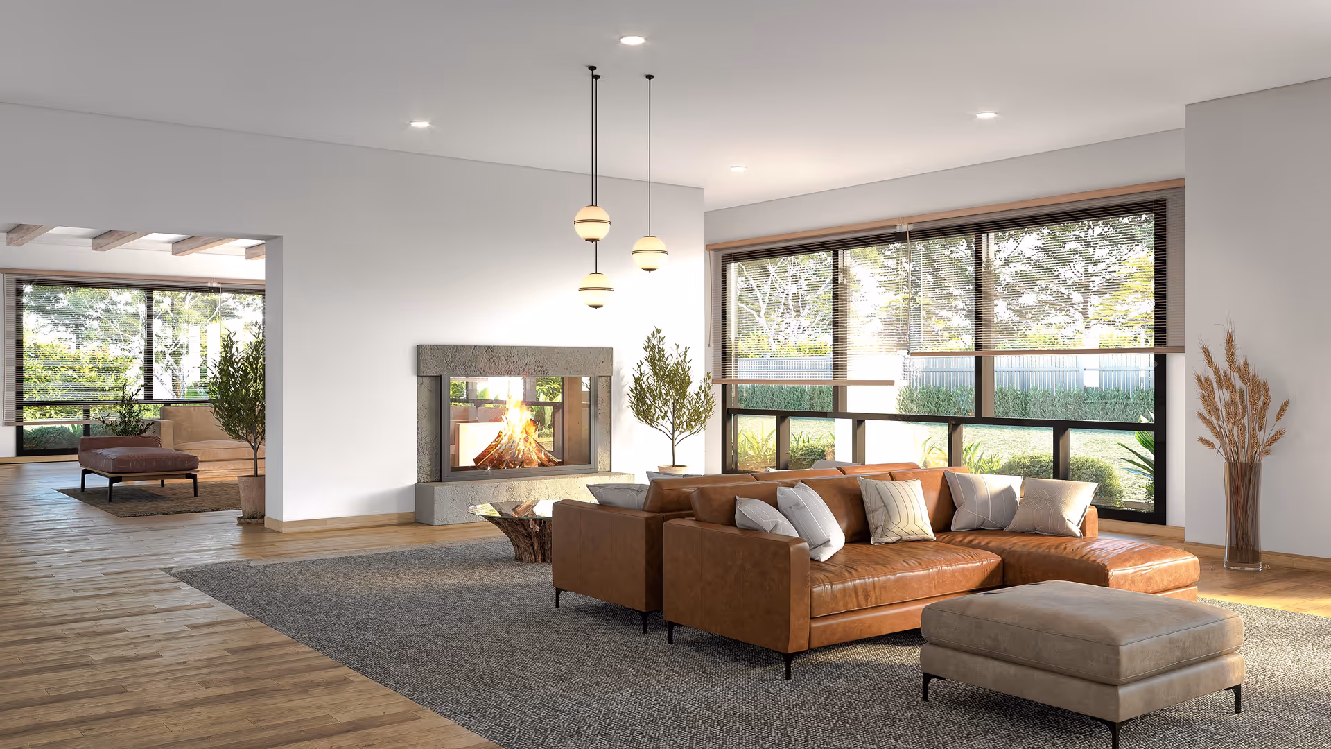 A modern living room with a brown leather sectional sofa adorned with multiple pillows, a beige ottoman, a textured gray area rug, and a glass coffee table with a wooden base. The room features a large window with blinds, a stone fireplace with a fire burning, three hanging pendant lights, and decorative plants. Adjacent to the living room is another seating area with a brown ottoman and a beige armchair near large windows showing greenery outside.