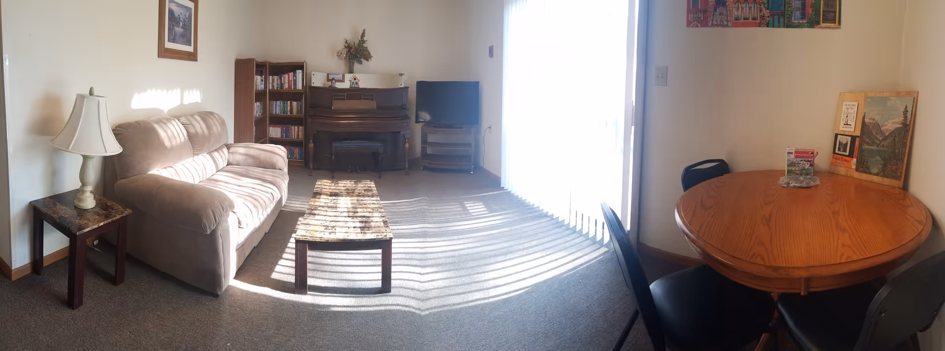 Bright living room with beige sofa, coffee table, piano, bookshelf, and round dining table.
