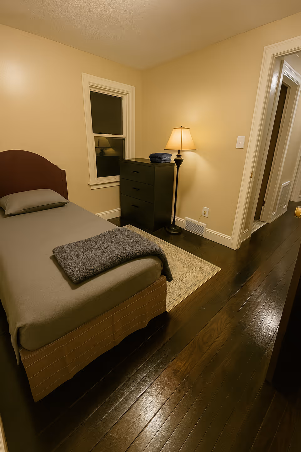 Cozy bedroom in a senior care home with bed, dresser, lamp, and dark wood floor.