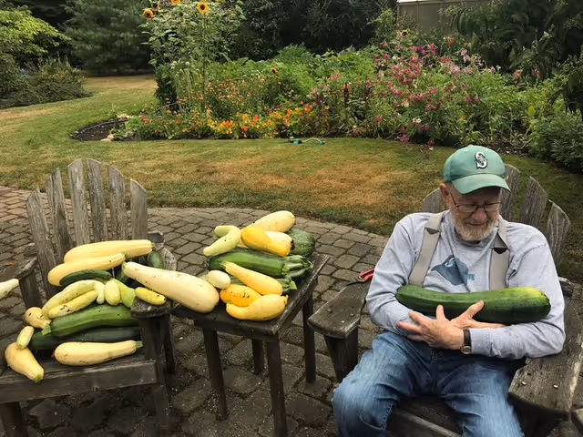 An elderly man sits outdoors with a table of yellow and green squash.